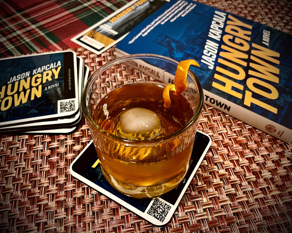 Glass of amber liquid with an ice sphere and orange twist, sitting next to Hungry Town coasters and book