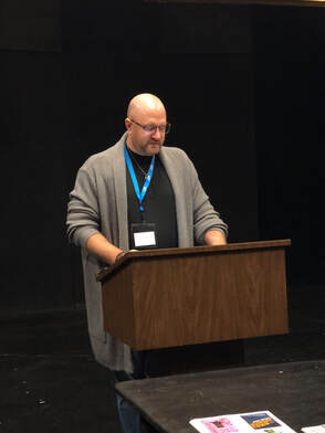 Jason Kapcala standing at a podium in a dark theatre, reading